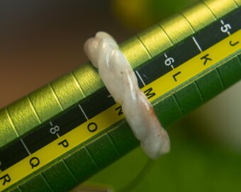May include: A pale, translucent, off-white ring with a twisted, rope-like texture. The ring is positioned on a green, cylindrical ring sizer tool, resting between the size markers M and N on the printed scale.