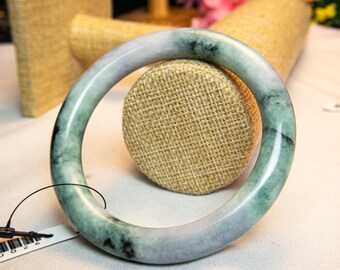 Op de afbeelding: Een jade armband met een glad, gepolijst oppervlak. De armband heeft een mix van groene, grijze en zwarte tinten. Het cirkelvormige ontwerp wordt gepresenteerd tegen een neutrale achtergrond, waarbij de kleurvariaties van de natuursteen worden benadrukt.