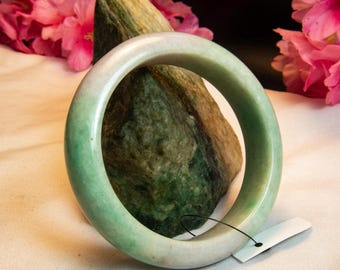 Op de afbeelding: Een jade armband met een glad, gepolijst oppervlak. De armband heeft een kleurovergang, van een bleek, crèmig wit naar een levendig groen. De armband wordt getoond op een getextureerde groene steen, met roze bloemen op de achtergrond.