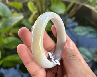 May include: A smooth, polished, translucent light-colored bangle bracelet with subtle yellow and white marbled tones. The circular jewelry piece features a small white rectangular tag attached with a thin black string, displaying a barcode.