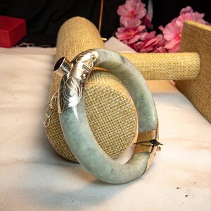 May include: A light green jade bangle bracelet with silver accents and a purple gemstone. The bracelet is displayed on a neutral-toned surface, showcasing its smooth, polished texture and intricate design.