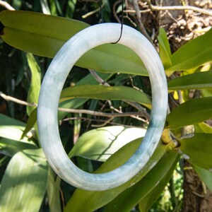 Könnte beinhalten: Ein hellgrünes Jade-Armreif-Armband. Das Armband hat eine solide, kreisförmige Form mit glatter Oberfläche. Der Jade hat eine durchscheinende Qualität mit Farbvariationen von hell- bis dunkelgrün. Das Armband hängt vor einem Hintergrund aus grünen Blättern.