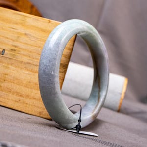 May include: A smooth, polished jade bangle bracelet featuring a rounded profile with a translucent, pale green and grey mottled appearance. The circular jewelry piece has a glossy finish and includes a thin black string tag attached to the bottom.
