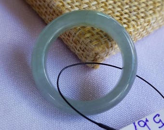 May include: A pale green jade bangle bracelet. The smooth, circular bracelet is displayed against a textured beige background and a light purple surface. A black string is attached to the bracelet. The bracelet is a classic jewelry piece.