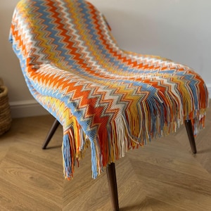 May include: A multi-coloured chevron patterned throw blanket draped over a chair. The blanket features a vibrant mix of orange, yellow, blue, and white, with a fringed edge. The chair has dark wooden legs.