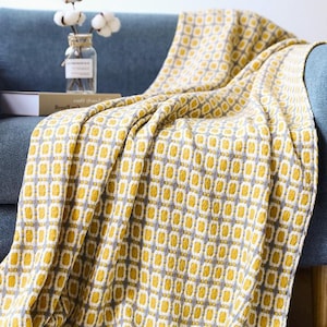 Op de afbeelding: Een gele en grijze deken met een geometrisch patroon. De deken is gemaakt van een zachte, geweven stof en heeft een gefranst randje.