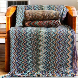 May include: A blue, green and brown chevron patterned throw blanket with fringe on the bottom edge. The blanket is folded and draped over a wooden armchair.