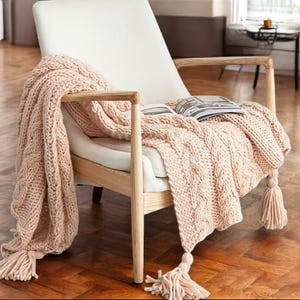 Puede incluir: Una manta de punto trenzado rosa claro con borlas sobre un sillón blanco con brazos y patas de madera. Una revista descansa sobre la manta. Un elemento de decoración hogareña acogedor.