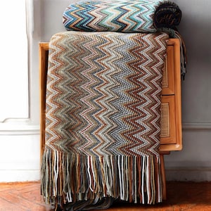 May include: A brown and blue chevron patterned throw blanket with fringe. The blanket is folded and draped over a wooden side table.