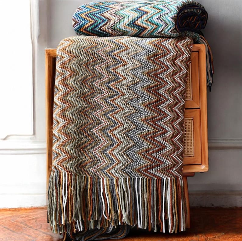May include: A brown and blue chevron patterned throw blanket with fringe. The blanket is folded and draped over a wooden side table.