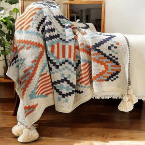 May include: A white throw blanket with a geometric pattern in shades of orange, blue, and black. The blanket has a fringe border and tassels.