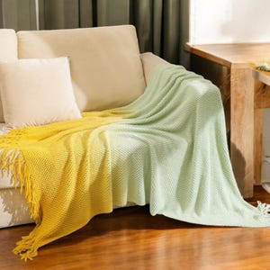 May include: A yellow and green ombre throw blanket with fringe on the edges. The blanket is draped over a sofa.