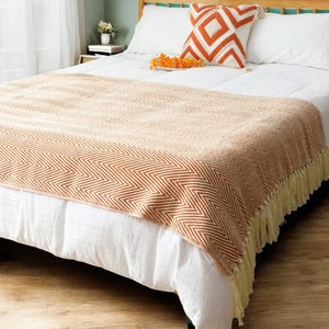 Puede incluir: Una suave manta de color melocotón con un patrón de chevron y ribete de flecos. La manta está colocada sobre una cama blanca con una almohada blanca y una almohada decorativa con un patrón geométrico naranja y blanco.