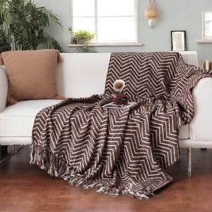 May include: A brown and white chevron patterned throw blanket with fringe, draped over a white sofa. A matching brown pillow with tassels rests on the sofa. A small decorative gramophone sits on the blanket.