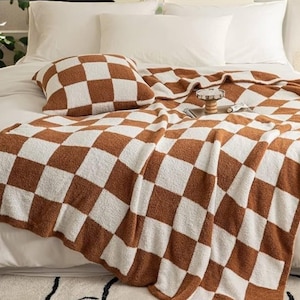 Könnte beinhalten: Ein braun-weiß kariertes Decken- und Kissen-Set auf einem Bett mit weißer Bettwäsche. Die Decke ist über das Bett drapiert, und das Kissen liegt darauf. Das Muster ist ein großes Schachbrettmuster.