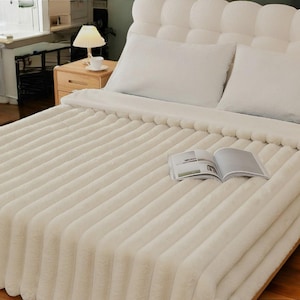 Op de afbeelding: Een wit, pluche bedsprei met een textuur, geribbeld ontwerp. De bedsprei is teruggevouwen om een wit laken en twee witte kussens te laten zien.