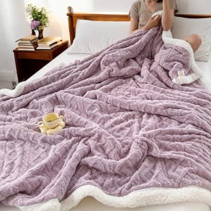 Pode incluir: Um cobertor macio, cor de lavanda, com um design de malha de cabo. O cobertor tem um forro e acabamento em sherpa branco macio. Uma pequena xícara de chá e pires decorativos estão sobre o cobertor. O cobertor está em uma cama com cabeceira de madeira.