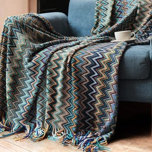 Puede incluir: Una manta de tiro de chevron azul y marrón con flecos en el borde inferior. La manta está drapeada sobre un sillón azul.