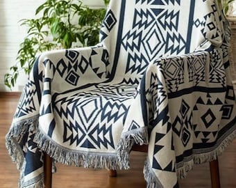 May include: A cream-colored throw blanket with a geometric navy blue pattern draped over a wooden chair. The blanket features diamond and zigzag designs and a fringed edge. The chair is set against a backdrop of greenery.