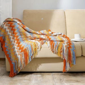 May include: A colourful throw blanket with a chevron pattern and fringe. The blanket is draped over a beige sofa.