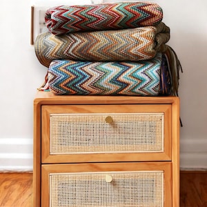 May include: A wooden bedside table with two drawers featuring woven cane fronts and brass knobs. A stack of colourful chevron patterned blankets sits on top of the bedside table.
