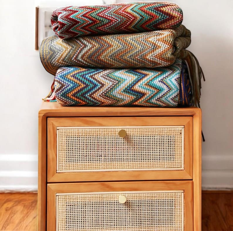 May include: A wooden bedside table with two drawers featuring woven cane fronts and brass knobs. A stack of colourful chevron patterned blankets sits on top of the bedside table.