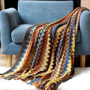 May include: A colourful knitted throw blanket with a chevron pattern in shades of brown, yellow, blue, and orange. The blanket has fringe on the edges and is draped over a blue armchair.