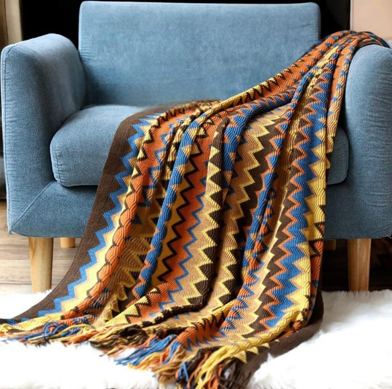 May include: A colourful knitted throw blanket with a chevron pattern in shades of brown, yellow, blue, and orange. The blanket has fringe on the edges and is draped over a blue armchair.