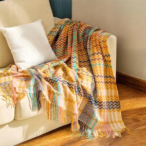 May include: A patterned throw blanket in shades of yellow, orange, and blue is draped over a cream sofa. The blanket features a chevron design and fringed edges with pink, green, and yellow tassels. A white cushion rests on the sofa.