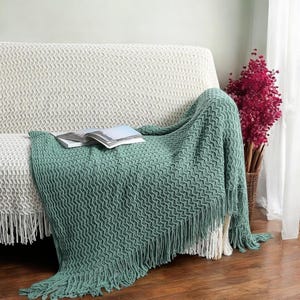 Puede incluir: Una manta de tejido de punto verde salvia con flecos, sobre un sofá blanco. La manta tiene un patrón de ondas texturizado. Un libro descansa sobre la manta. El sofá está cubierto con una funda de punto blanca.