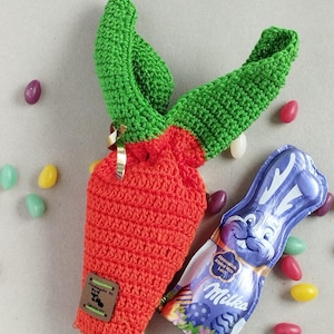 May include: A handmade crochet carrot-shaped gift bag in bright orange with green leaves. A small tag reads "Handmade by..." and a foil-wrapped chocolate bunny is next to the bag. Colorful jelly beans are scattered around the items.
