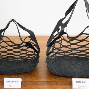 May include: Two black tote bags with unique cut-out designs. The bags have a net-like pattern and sturdy handles. One is labeled "Market Bag" and the other "Tote Bag". They are displayed on a wooden surface.