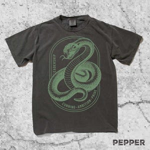 May include: Dark gray t-shirt with a green snake graphic inside an oval frame. The frame includes the words "Leadership", "Cunning - Ambition - Pride". The word "Pepper" is printed at the bottom right.