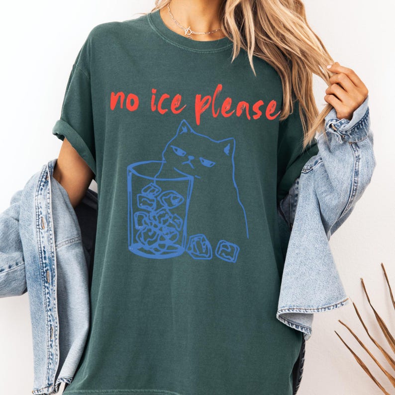 K&ouml;nnte beinhalten: Dunkelgr&uuml;nes T-Shirt mit dem roten Text "no ice please". Eine blaue Illustration einer m&uuml;rrischen Katze und eines Glases Eisw&uuml;rfel ist auf der Vorderseite aufgedruckt. Das T-Shirt hat eine lockere Passform und kurze &Auml;rmel.
