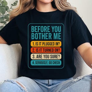 Peut inclure: T-shirt noir avec le texte "BEFORE YOU BOTHER ME" en turquoise, avec une liste de questions en blanc et orange. Le design comprend les phrases : "IS IT PLUGGED IN ?", "IS IT TURNED ON ?", "ARE YOU SURE ?" et "SERIOUSLY, GO CHECK."