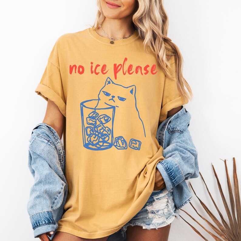 K&ouml;nnte beinhalten: Senfgelbes T-Shirt mit dem roten Text "no ice please". Eine blaue Umrisszeichnung einer grummeligen Katze befindet sich neben einem Glas Eisw&uuml;rfeln. Das T-Shirt hat hochgekrempelte &Auml;rmel.