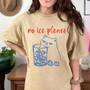K&ouml;nnte beinhalten: Beigefarbenes T-Shirt mit dem roten Text "no ice please". Eine blaue Illustration einer m&uuml;rrischen Katze befindet sich neben einem Glas Eisw&uuml;rfeln. Das T-Shirt hat einen l&auml;ssigen Stil.