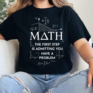 May include: Black t-shirt featuring white text and mathematical symbols. The word "MATH" is central, with the phrase "THE FIRST STEP IS ADMITTING YOU HAVE A PROBLEM" below. The design also includes various equations and graphs.