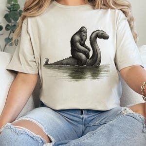 Peut inclure: T-shirt beige avec une illustration en noir et blanc de Bigfoot chevauchant le monstre du Loch Ness dans l'eau. Le graphisme est un dessin de style vintage. Le t-shirt a un col rond et des manches courtes.