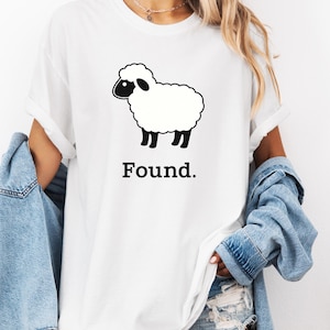 Puede incluir: Camiseta blanca con un gráfico de oveja de dibujos animados en blanco y negro. La oveja tiene un cuerpo esponjoso, cara y patas negras. La palabra "Found." está impresa debajo de la oveja en negrita, en negro. La camiseta es de estilo casual.