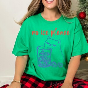 K&ouml;nnte beinhalten: Gr&uuml;nes T-Shirt mit dem roten Text "no ice please". Das Shirt zeigt eine blaue Illustration einer grummeligen Katze neben einem Glas Eis. Das Shirt ist ein l&auml;ssiger, bequemer Stil.
