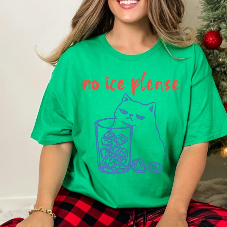 K&ouml;nnte beinhalten: Gr&uuml;nes T-Shirt mit dem roten Text "no ice please". Das Shirt zeigt eine blaue Illustration einer grummeligen Katze neben einem Glas Eis. Das Shirt ist ein l&auml;ssiger, bequemer Stil.