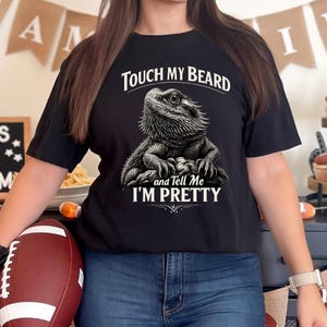 Peut inclure: T-shirt noir avec une illustration détaillée d'un dragon barbu et le texte "Touch My Beard and Tell Me I'm Pretty" en blanc. Le graphisme est centré sur le t-shirt. Le t-shirt est fait d'une matière douce.