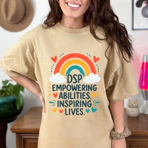 May include: A beige t-shirt featuring a rainbow design and the words "DSP EMPOWERING ABILITIES. INSPIRING LIVES." The design incorporates hearts and clouds. The shirt is made of a soft material.