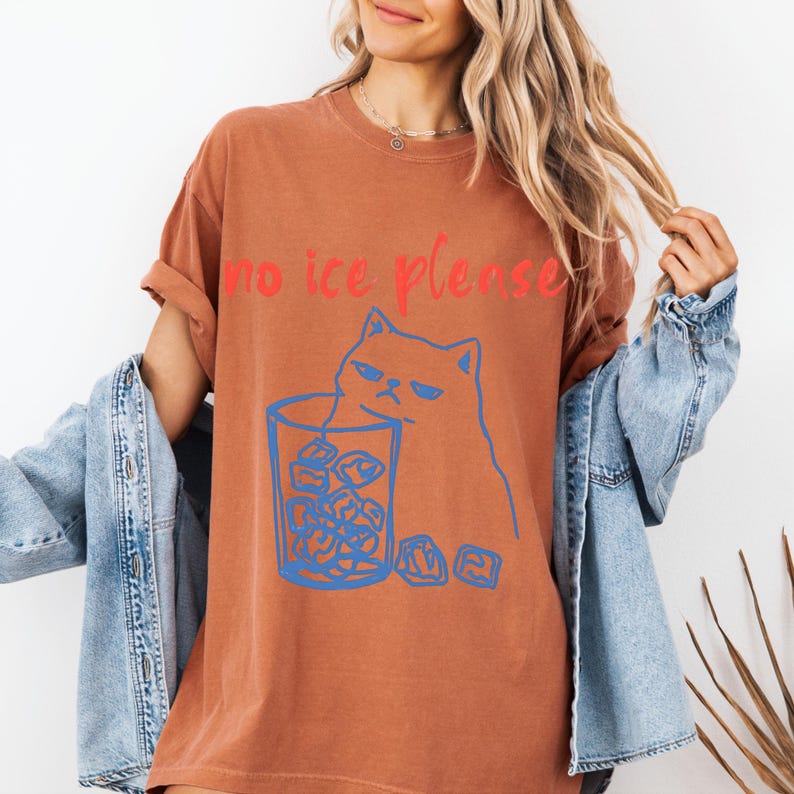 K&ouml;nnte beinhalten: Rostfarbenes T-Shirt mit dem roten Text "no ice please". Eine blaue Illustration einer m&uuml;rrischen Katze befindet sich neben einem Glas Eisw&uuml;rfeln. Das T-Shirt hat hochgekrempelte &Auml;rmel.