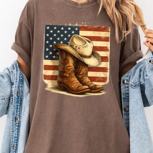Peut inclure: T-shirt marron avec un motif de chapeau et de bottes de cowboy devant un drapeau américain vieilli. Les bottes sont marron et le chapeau est beige. Le drapeau a des rayures rouges et blanches et des étoiles bleues.