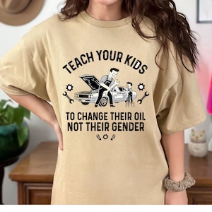 May include: Tan t-shirt with the text "TEACH YOUR KIDS TO CHANGE THEIR OIL NOT THEIR GENDER" above a graphic of a car being worked on by a parent and child. The design includes wrenches and gears.