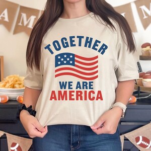 Peut inclure: Un t-shirt beige avec le texte "TOGETHER WE ARE AMERICA" en bleu et rouge, orné d'un drapeau américain. Le t-shirt est à manches courtes. L'arrière-plan comprend des bannières décoratives et des aliments.