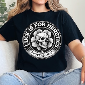 Peut inclure: T-shirt noir avec un motif circulaire blanc. Le motif représente un crâne dans une fleur, avec le texte "LUCK IS FOR HERETICS" et "PROVERBS 16:55". Le t-shirt est un vêtement décontracté.