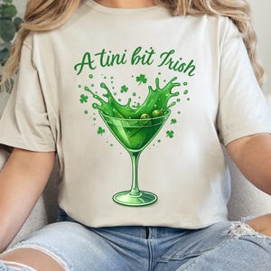 Könnte beinhalten: Beiges T-Shirt mit einem grünen Martini-Glas-Motiv. Die Grafik zeigt einen Spritzer grüne Flüssigkeit, Oliven und Kleeblätter, mit dem Text "A tini bit Irish" in grüner Schrift. Das T-Shirt ist für Freizeitkleidung geeignet.